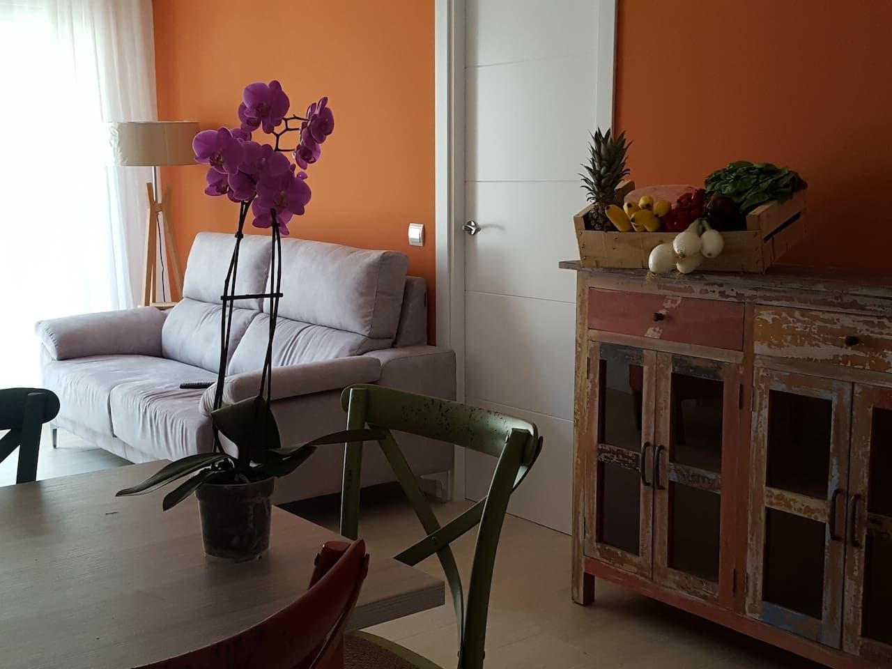 A living room with a sofa, a dining table with chairs, a potted plant, and a cabinet with food items.