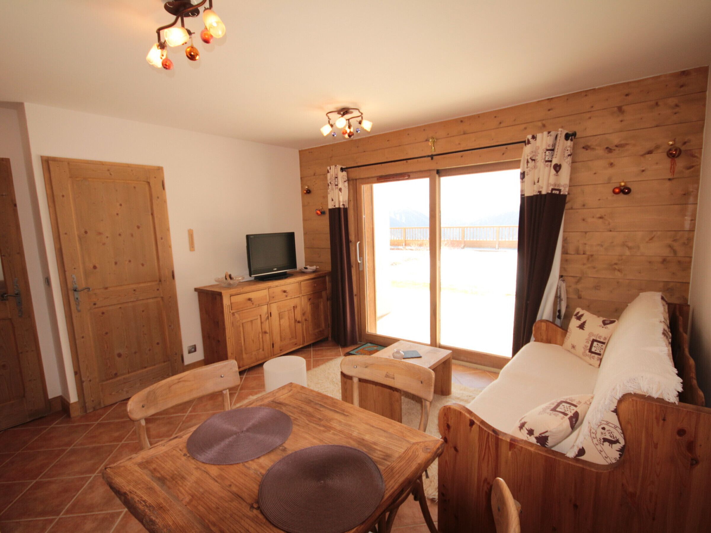 A cozy room with a wooden dining table, chairs, a sofa, a television, and a sliding glass door leading to a balcony.