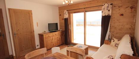 A cozy room with a wooden dining table, chairs, a sofa, a television, and a sliding glass door leading to a balcony.
