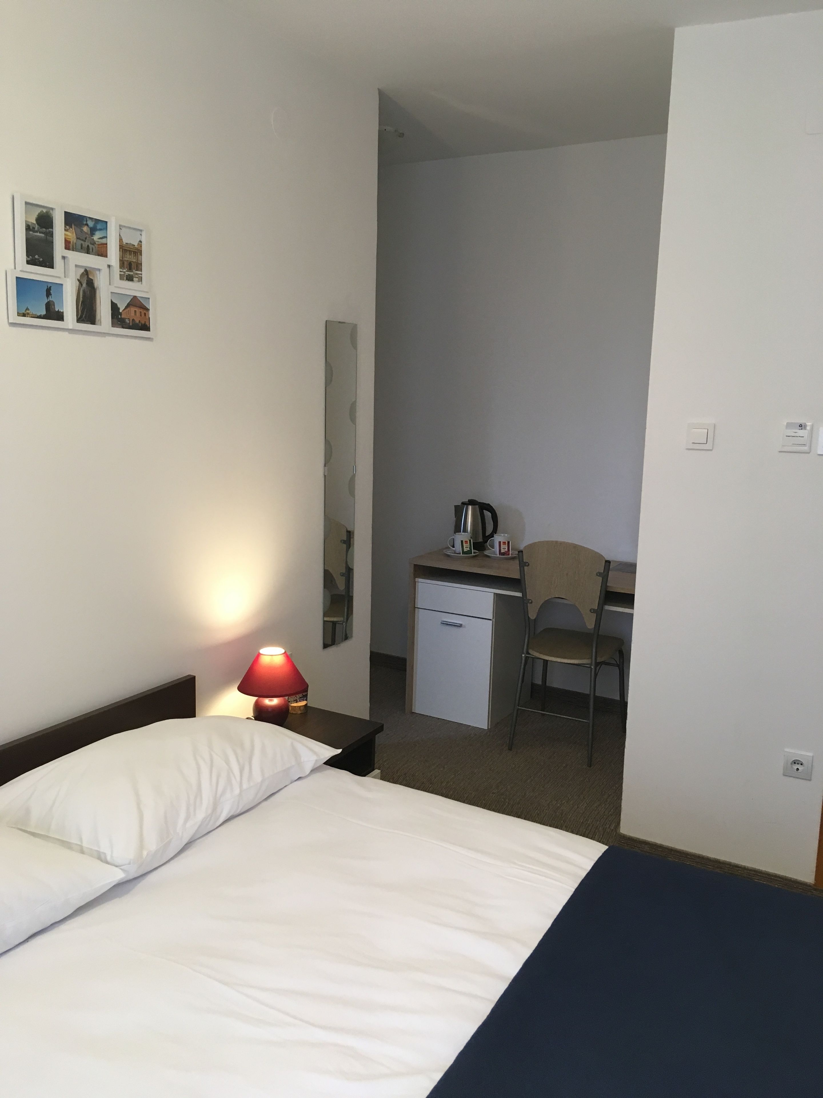 Hotellihuone, jossa on sänky, työpöytä tuolilla, pieni jääkaappi ja lamppu.