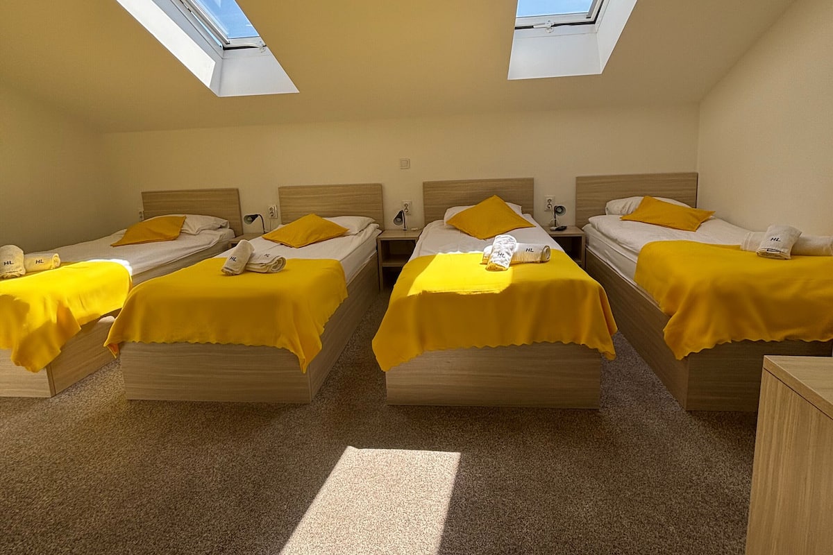 A room with four beds, each with a yellow bedspread and pillows, arranged in a U-shape. There are skylights on the ceiling, allowing natural light to enter.