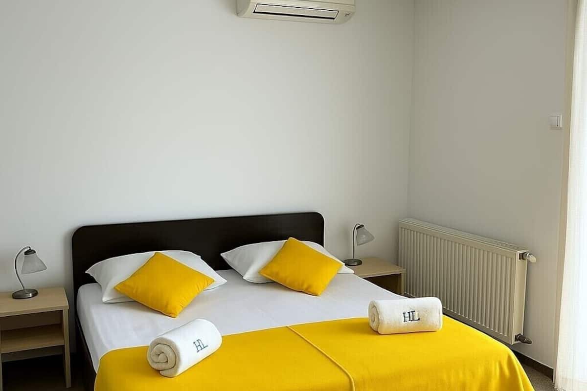A hotel room with a bed, two pillows, a nightstand, an air conditioning unit, a radiator, and a window with curtains.