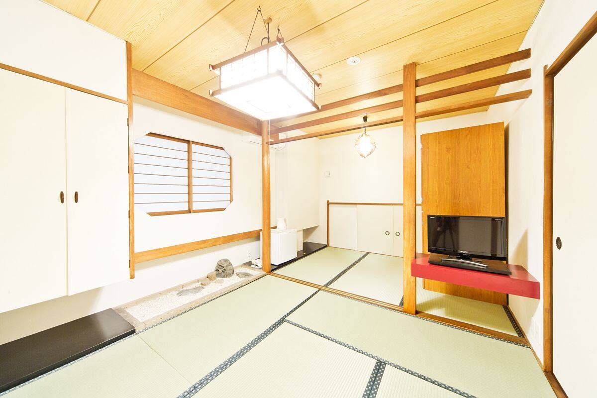Une pièce de style japonais traditionnel, avec un sol recouvert de tatamis, un téléviseur posé sur un meuble rouge et des portes coulissantes en bois.