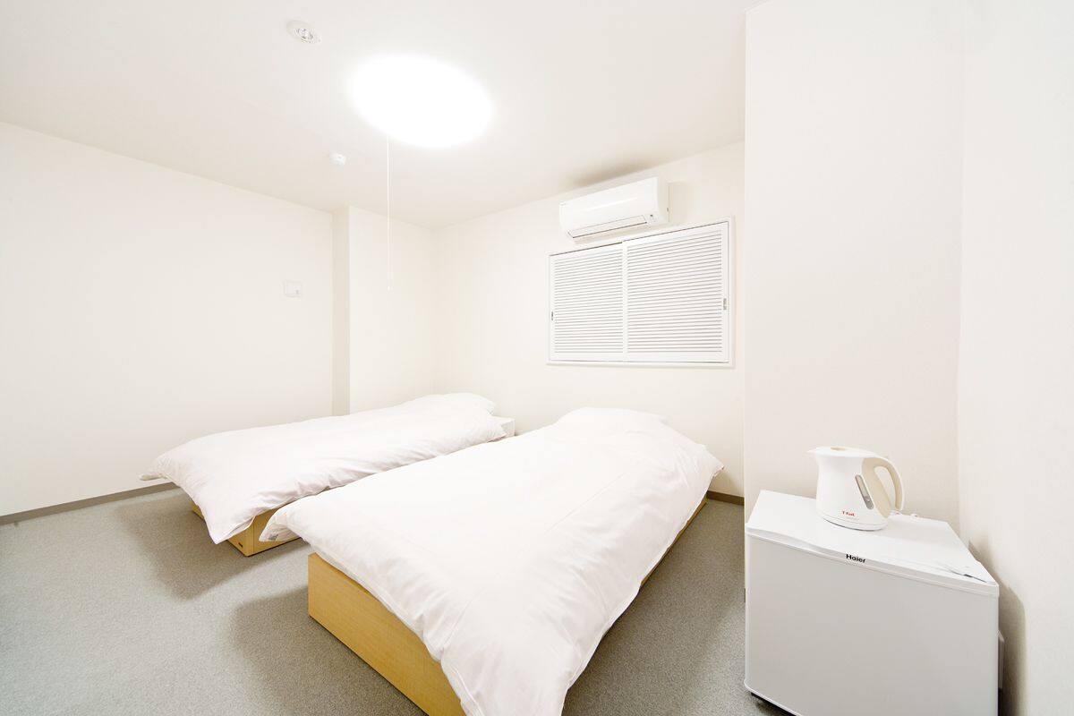 Une chambre d’hôtel minimaliste avec deux lits simples, un petit radiateur blanc et une bouilloire posée sur une table d’appoint.