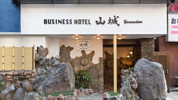 Entrada de un hotel de negocios con fachada de piedra, grandes rocas y una puerta tradicional japonesa.