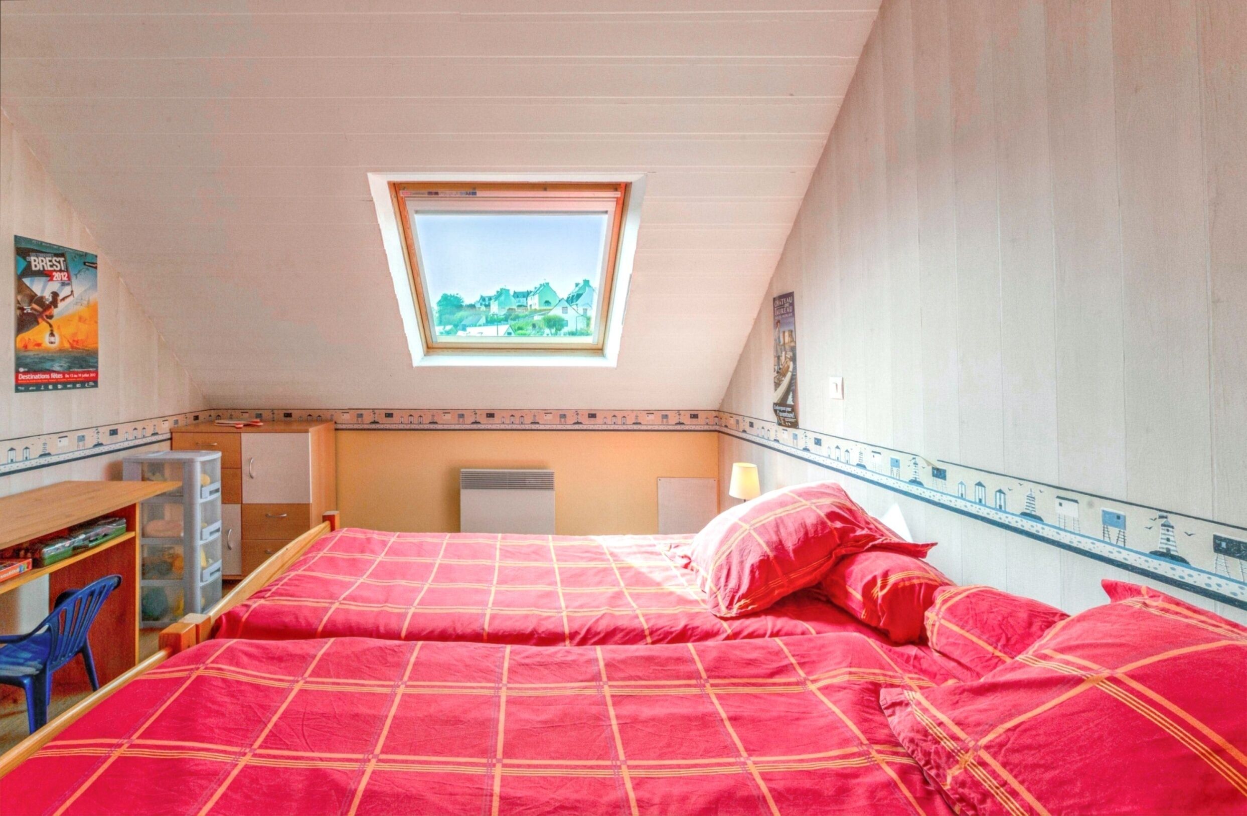 A bedroom with a skylight, a bed with red bedding, a desk with a chair, and a poster on the wall.