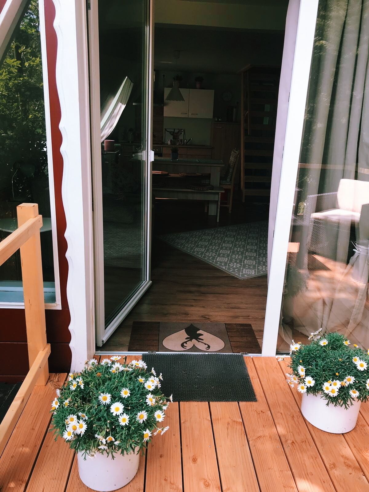 Een houten terras met twee potten margrietjes, een schuifdeur naar een kamer met een eettafel en stoelen, en een klein vloerkleed met een pootafdruk patroon.