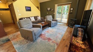 A living room with a sofa, armchair, and a television. There is a fireplace and a sliding glass door leading to a balcony. - Mountain Retreat at Lake Cushman (Hoodsport)