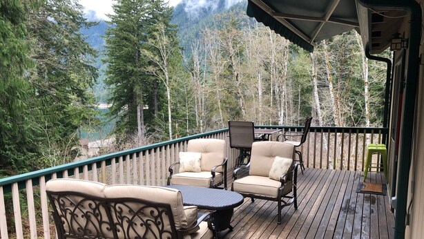 A wooden deck with outdoor furniture, including a sofa and chairs, overlooking a forested area with mountains in the background. - Mountain Retreat at Lake Cushman (Hoodsport)