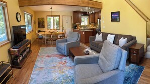 A cozy living room with a sofa, coffee table, and TV stand. There is a dining area with chairs and a table. The kitchen is visible in the background. - Mountain Retreat at Lake Cushman (Hoodsport)