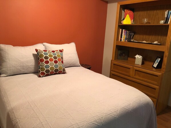 A neatly made bed with a white comforter and a colorful hexagonal pillow. A wooden nightstand with a lamp and books is beside the bed.