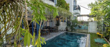 A swimming pool with a tiled deck, surrounded by greenery and a building with balconies.