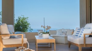Uma área externa iluminada pelo sol com vista para o mar, com uma mesa de madeira, duas cadeiras acolchoadas e uma cesta de vime.