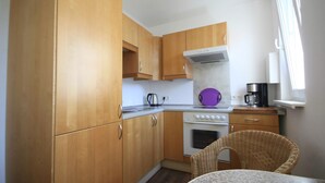 A compact kitchen with wooden cabinets, a stove, and a wicker chair. - Schöne 3Zimmer FeWo direkt am Baalensee (Fürstenberg-Havel)