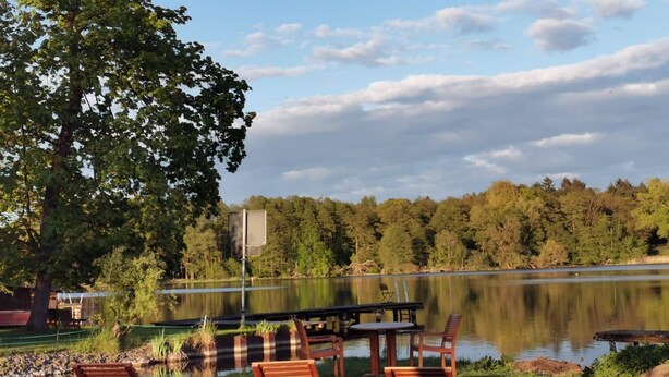 A lakeside outdoor seating area with wooden chairs and tables, surrounded by trees and a clear sky. - Schöne 3Zimmer FeWo direkt am Baalensee (Fürstenberg-Havel)