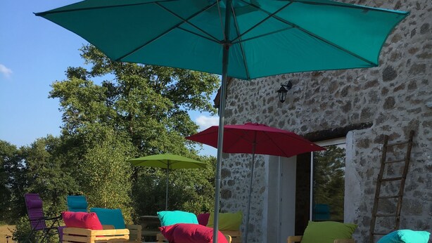 Outdoor seating with wooden crates and colorful cushions, a teal umbrella, and a stone building in the background.