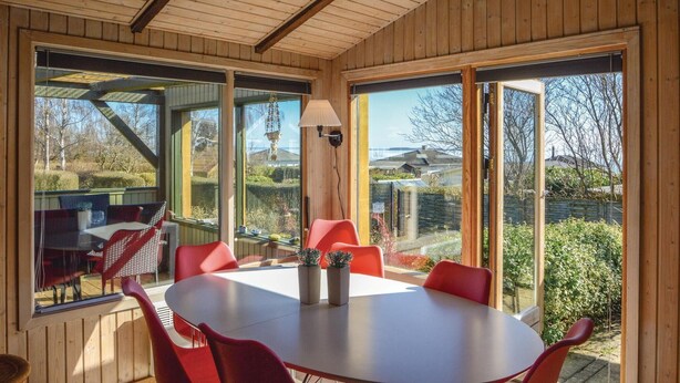 Een kamer met houten lambrisering, een ronde tafel en rode stoelen, grote ramen en uitzicht op een tuin en gebouwen.