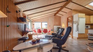 Een gezellig interieur van een chalet met een eethoek, een zithoek met een bank en stoelen, een keuken met een koelkast, en een houten plafond.