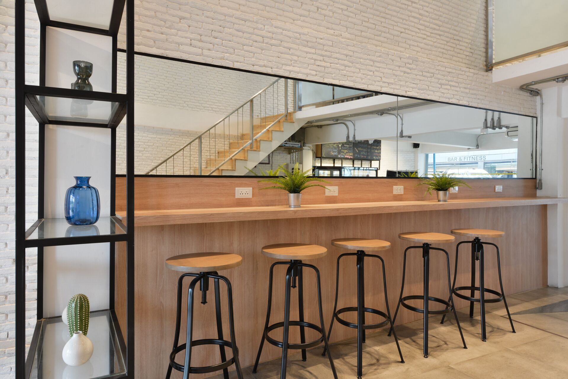 A modern bar area with a wooden counter, black metal stools, and a staircase in the background.