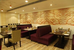 A restaurant with a high ceiling, recessed lighting, and a decorative wall with floral patterns. There are several tables and cushioned seating areas. - WJ Grand Hotel (Jalandhar)