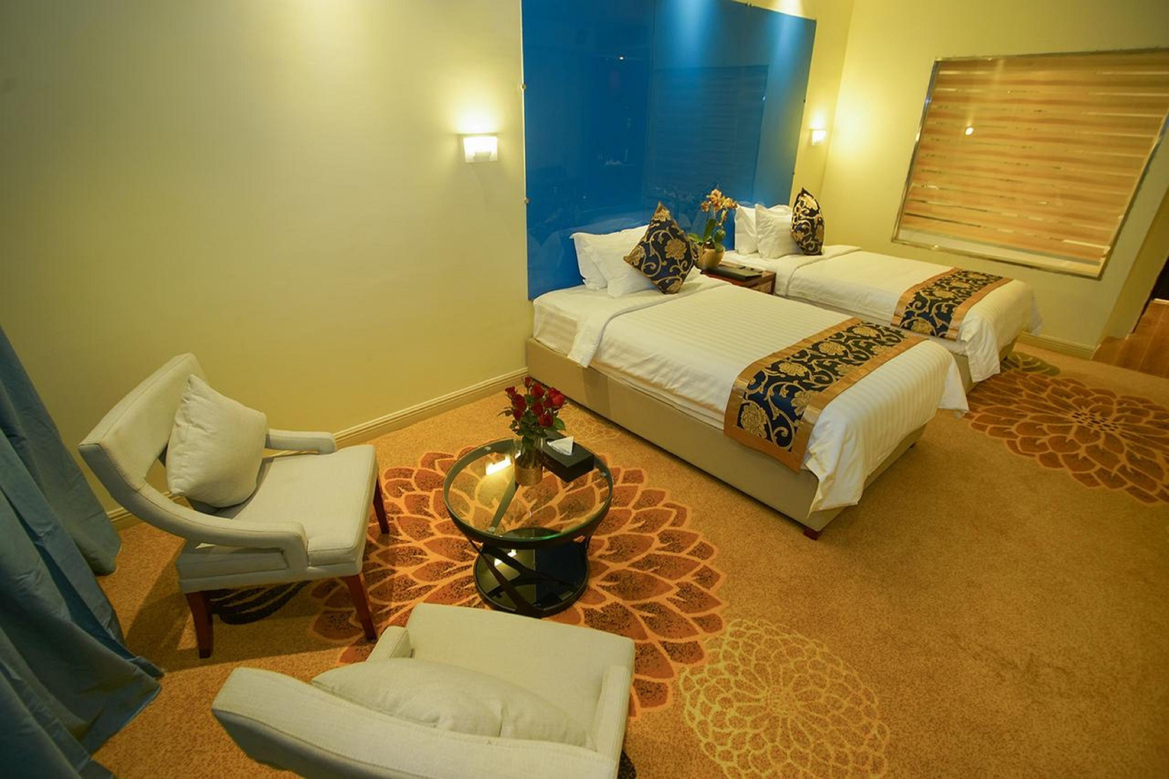A hotel room with two beds, a sitting area with a chair, a small table with a vase, and a patterned carpet.
