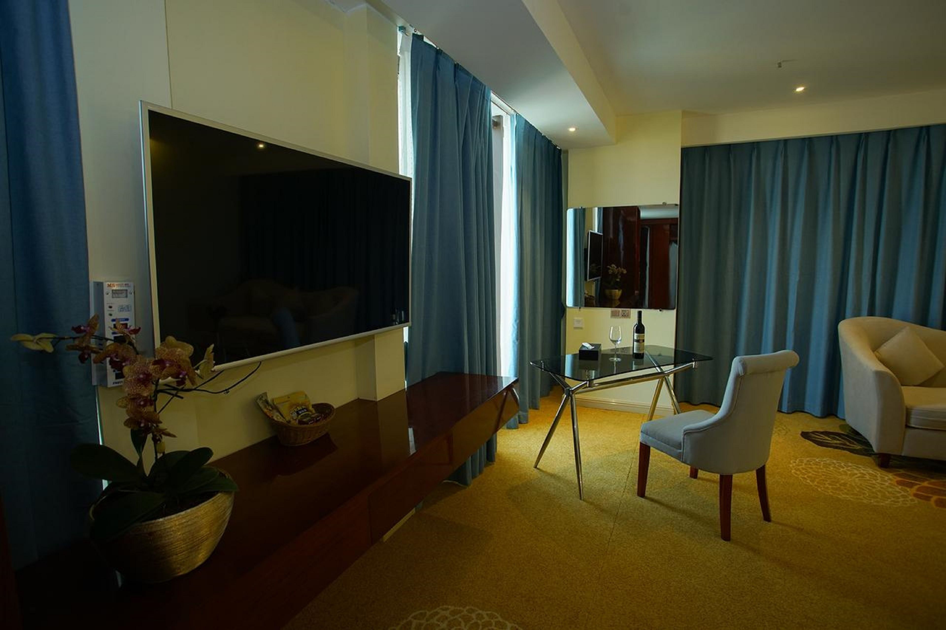 A hotel room with a large flat-screen TV, a dining area with a glass table, a sofa, and a chair.