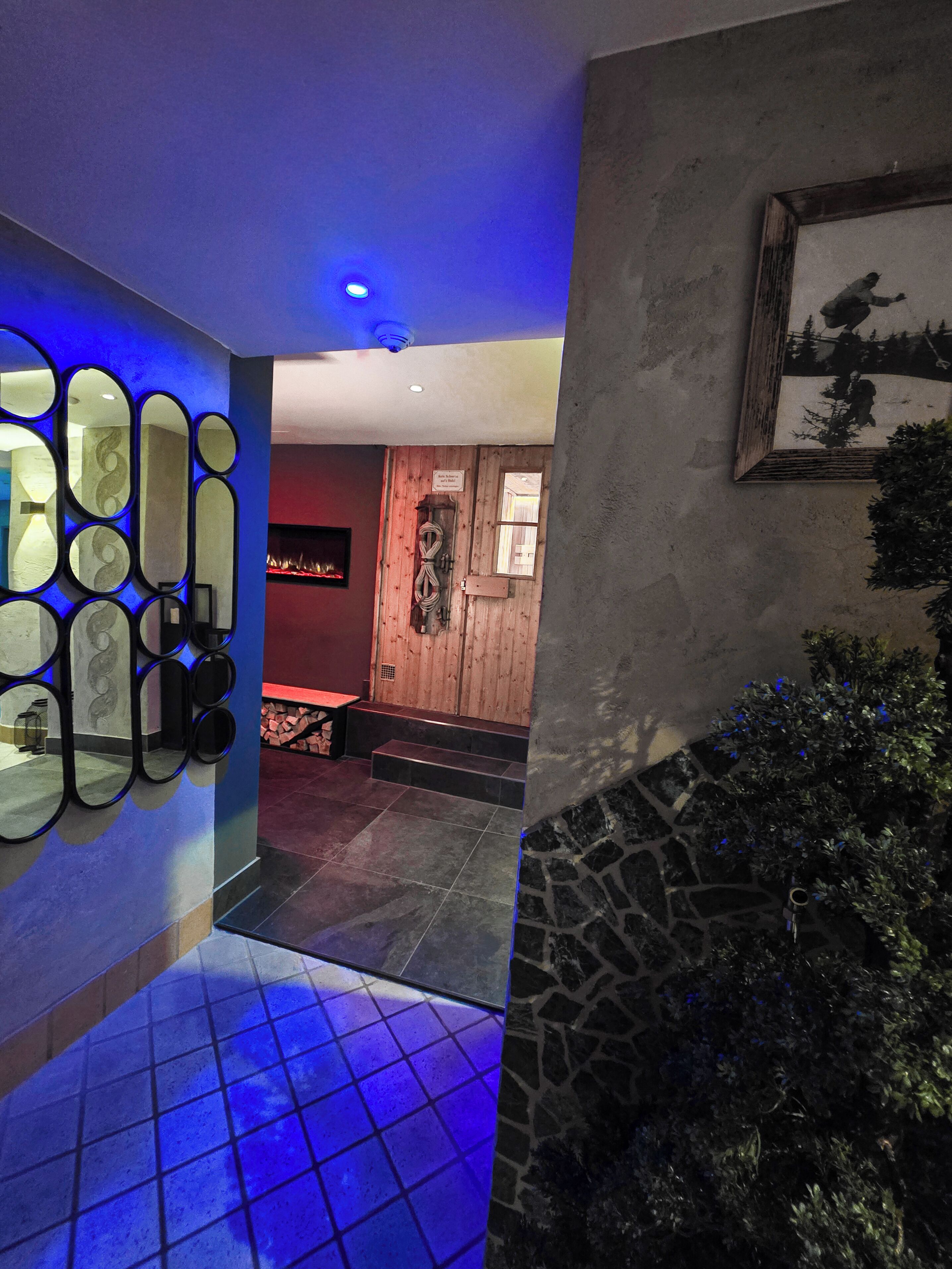 A hallway with a blue-lit wall, a stone wall with a framed picture, and a tiled floor.
