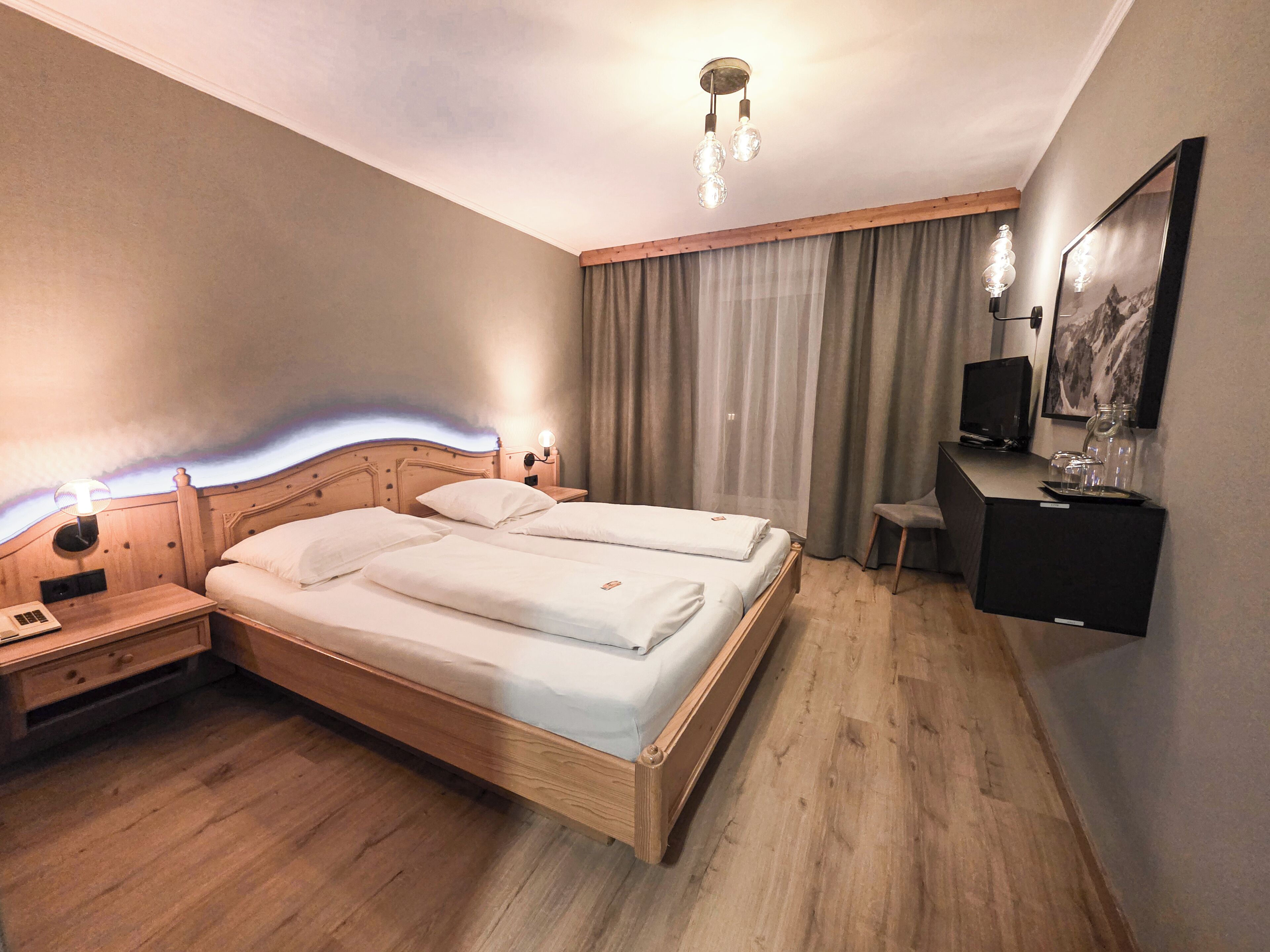 Hotellihuone, jossa on puinen sänky, työpöytä tuolilla, televisio ja taulu seinällä.