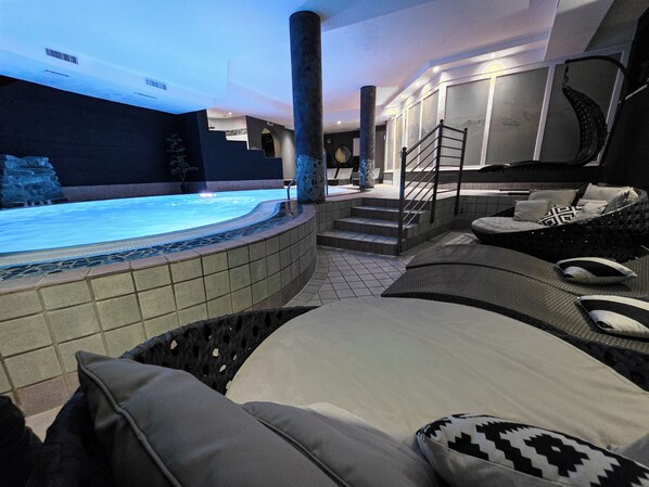 Un área de piscina interior moderna con un jacuzzi, zona de estar con cojines y una escalera que lleva a un nivel superior.