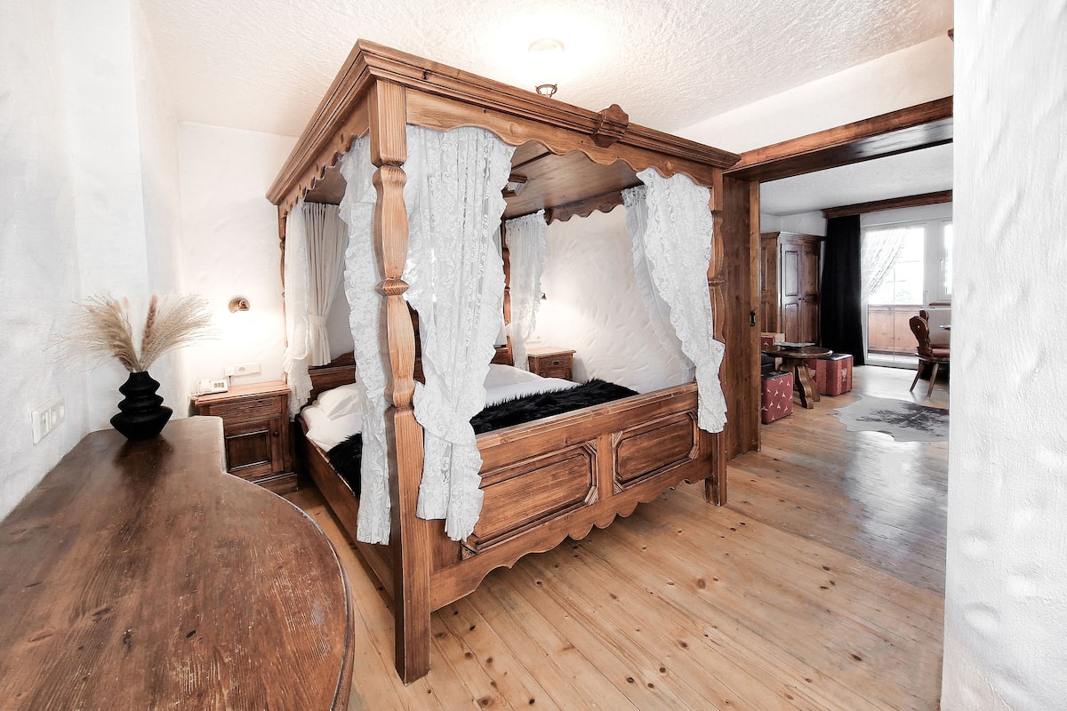 Una cama con dosel y cuatro columnas, con estructura de madera y cortinas blancas, un mesita de noche de madera y un florero con plumas de plumeros secos.