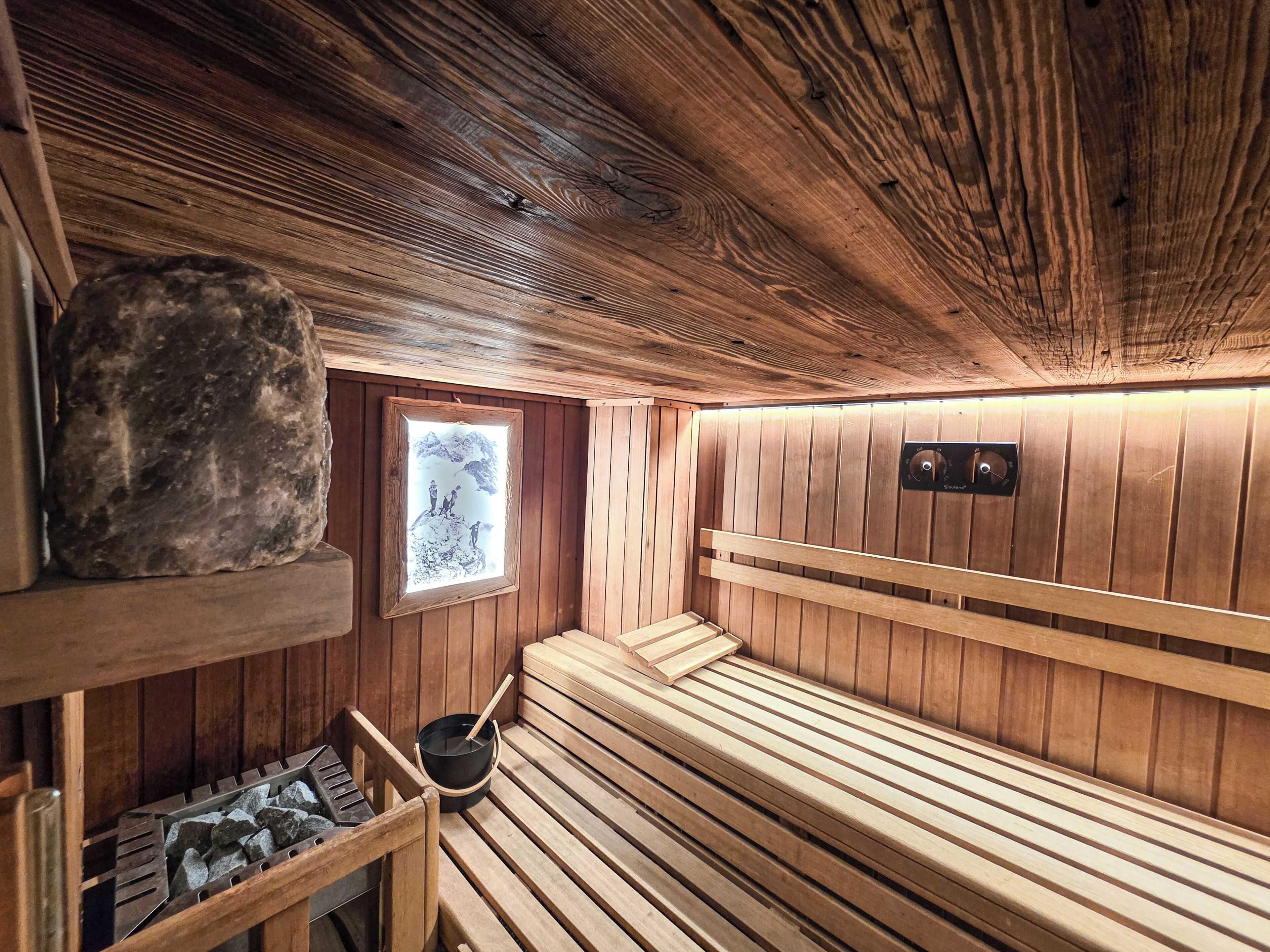 A sauna room with wooden benches, a stone shelf, a window with a view, and a control panel.