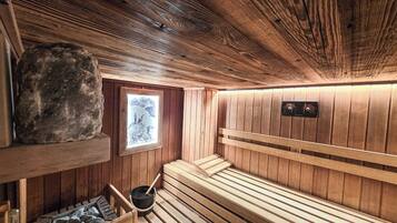 A sauna room with wooden benches, a stone shelf, a window with a view, and a control panel.