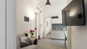 A modern living room with a sofa, a flat-screen TV mounted on the wall, and a kitchen area in the background. - La casa di Emanuela Guest house (Roma)