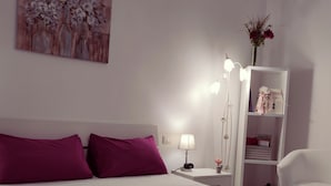 Een hotelkamer met een bed, nachtkastje, lamp, witte stoel, plank met spullen en een schilderij aan de muur.