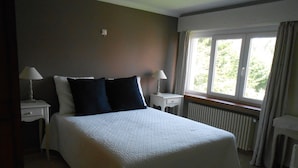 Un lit bien fait, avec une literie blanche et deux oreillers noirs, flanqué de deux tables de chevet sur lesquelles reposent des lampes, dans une pièce dotée d’une grande fenêtre et d’un radiateur.