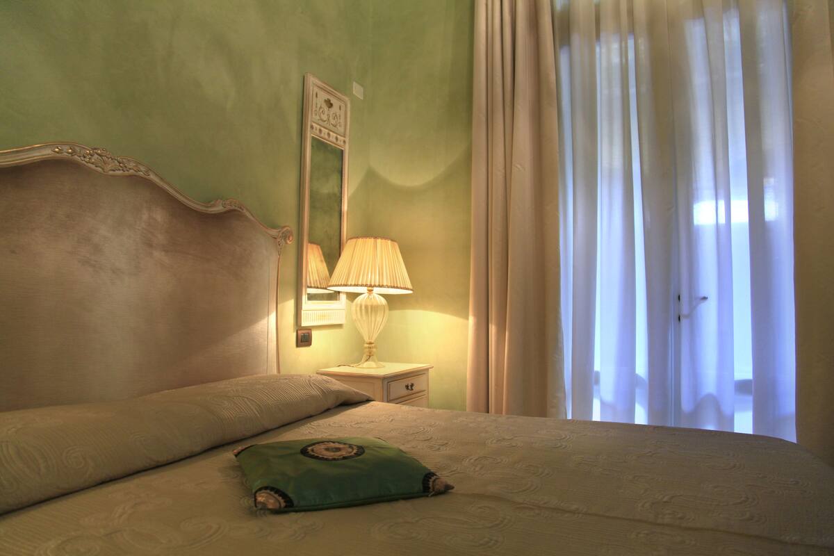 A neatly made bed with pillows, a bedside lamp, and a mirror on a nightstand.