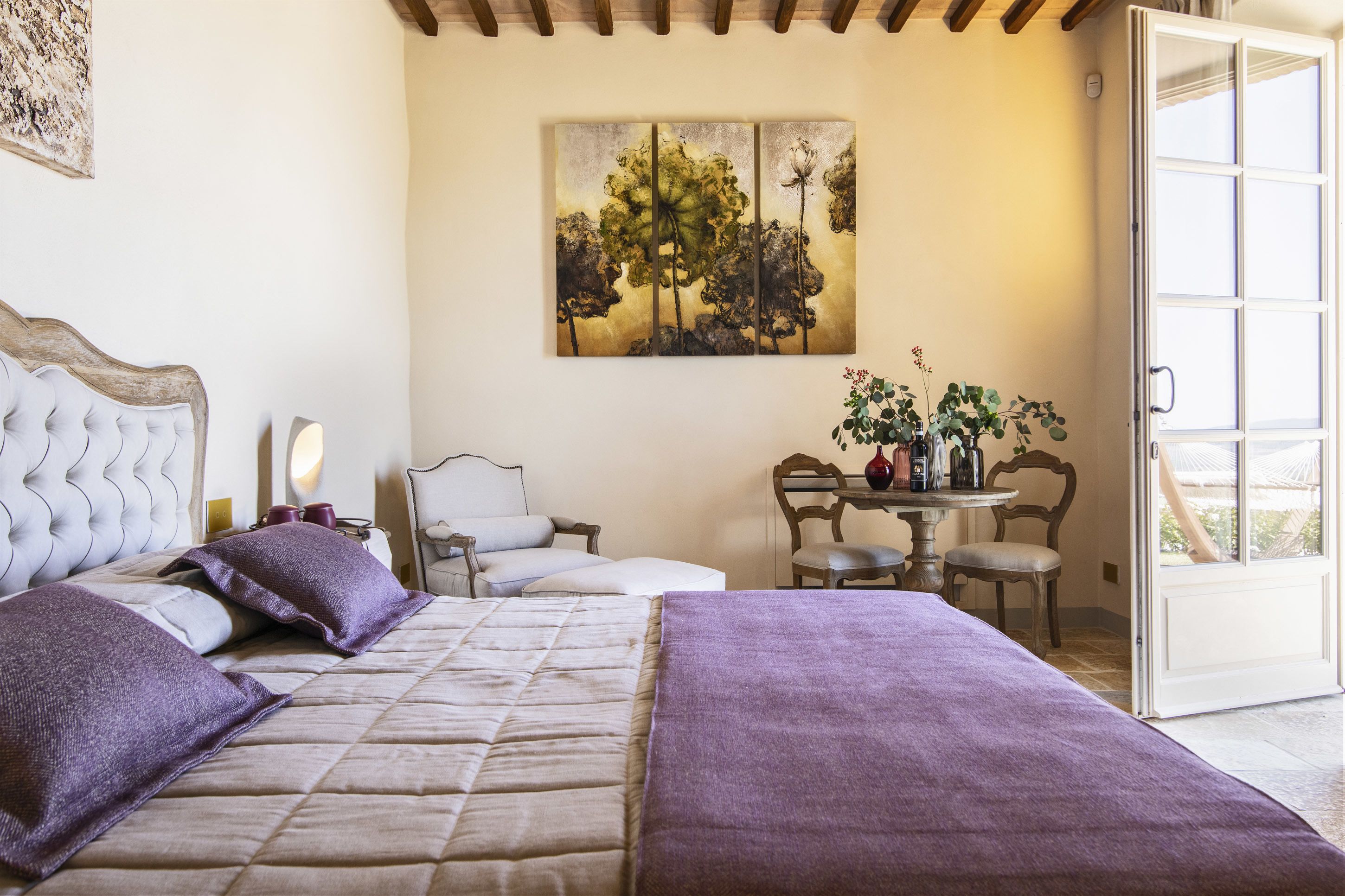 A bedroom with a bed, a chair, a table with a vase, and a painting on the wall.