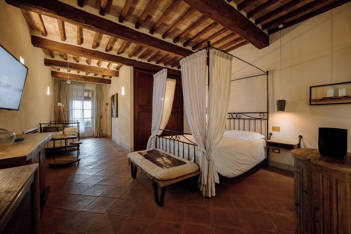 Une chambre à coucher avec un lit à baldaquin, un banc en bois, un bureau avec une chaise et un téléviseur posé sur une commode en bois.
