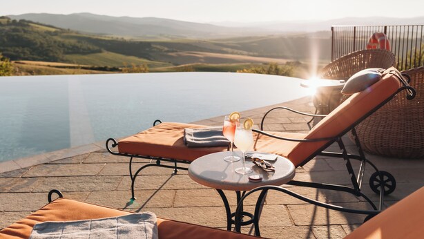 Zona junto a la piscina con sillas de descanso, una mesa con bebidas y vistas a colinas.