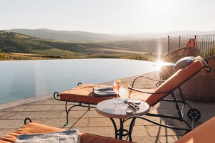 Zona junto a la piscina con sillas de descanso, una mesa con bebidas y vistas a colinas.