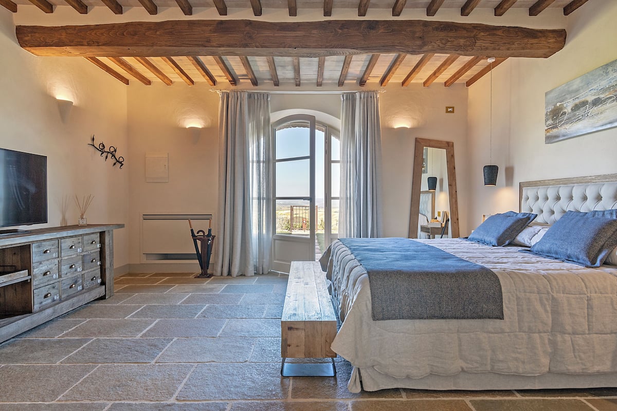 Une chambre avec un sol en pierre, un banc en bois, un lit avec une tête de lit, une télévision et une fenêtre avec des rideaux.
