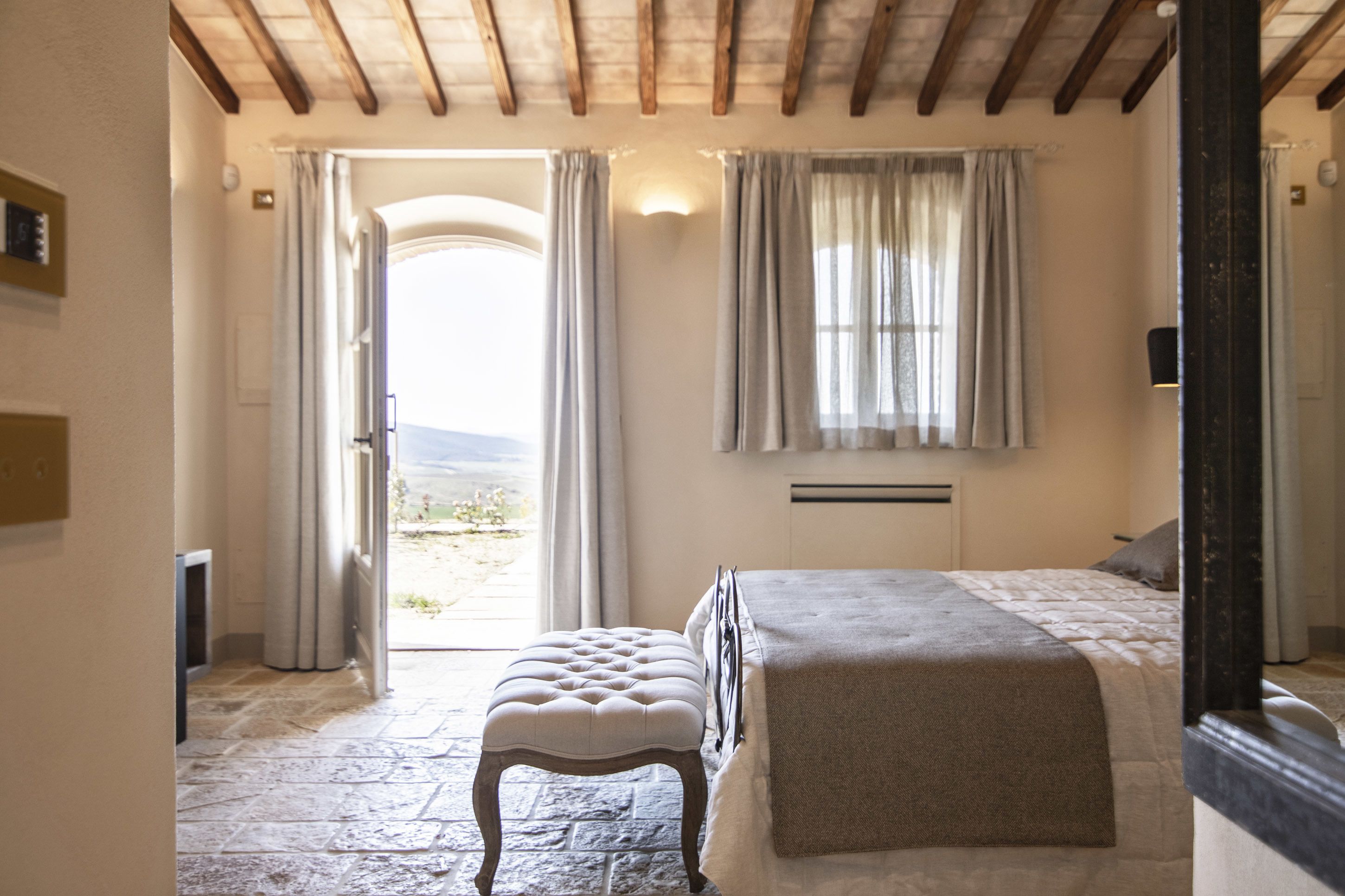 A bedroom with a stone floor, a bed with a grey blanket, a wooden bench, and a window with curtains.