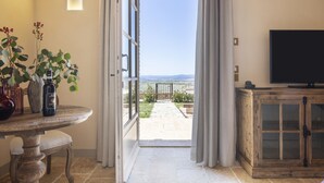 Puerta abierta que da a un patio con vistas al campo, una mesa con plantas y un televisor sobre un mueble de madera.