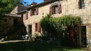 A stone building with red shutters, a garden with roses, and a clear blue sky.