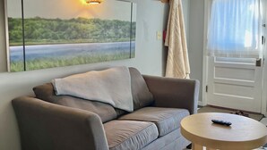 Una sala de estar con un sofá gris, una mesa redonda de madera, un cuadro de un paisaje, una lámpara colgante y una puerta con cortina.