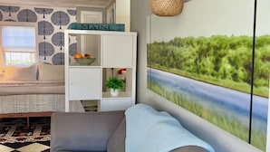Una sala de estar con un sofá gris, un mueble blanco con un frutero, una lámpara de pared y un cuadro de paisaje grande.