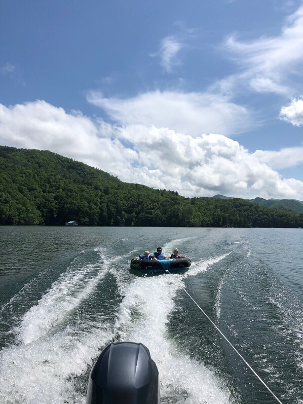 A watercraft towing a raft with passengers, a lake, and forested hills in the background.