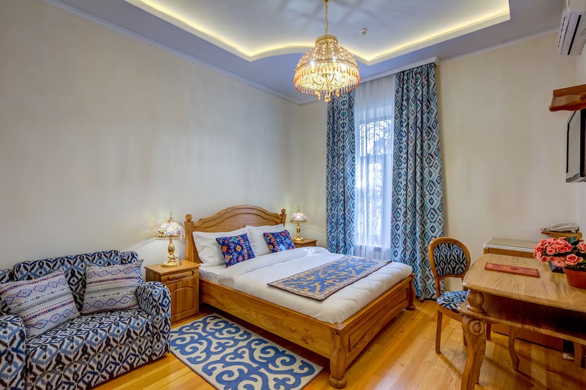 Ein Schlafzimmer mit einem hölzernen Bett, gemusterten Kissen, einem Nachttisch mit Lampe, einem blauen Teppich mit Muster und einem Esstisch mit Stühlen.