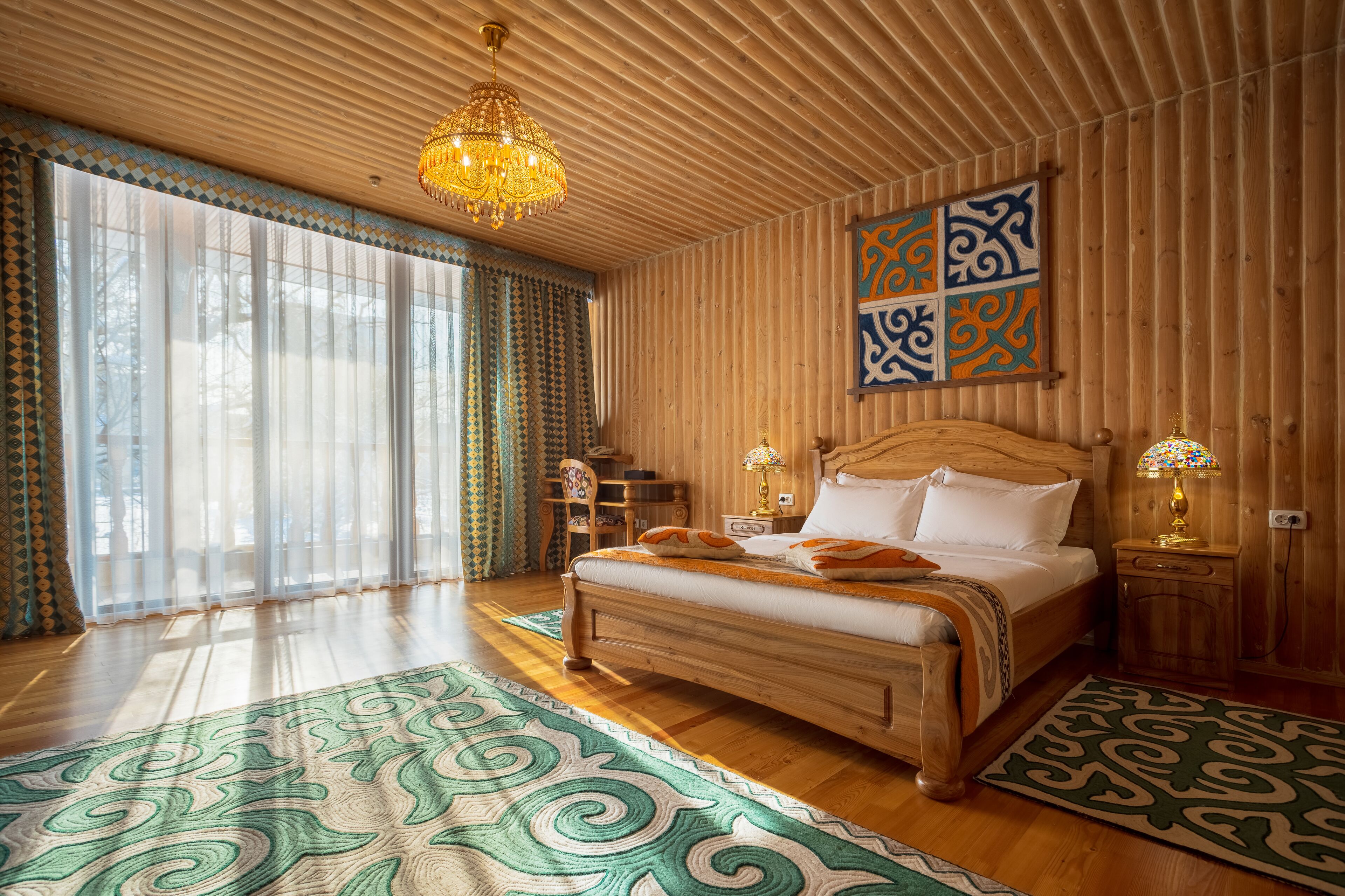 A wooden-paneled bedroom with a large bed, two bedside tables with lamps, a patterned rug, and a large window with sheer curtains.