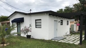 A small, single-story building with a white exterior and a dark blue awning. There is a potted plant with pink flowers in front of the entrance.